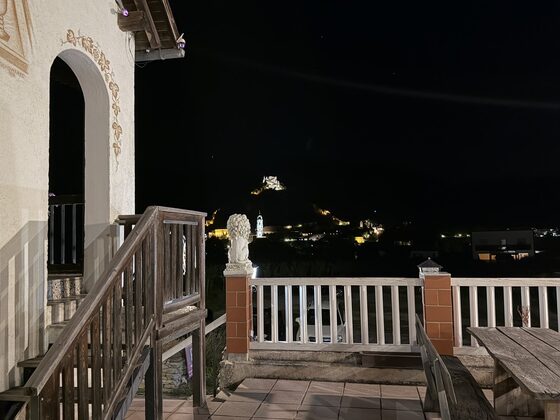 Holzstiege zur Terrasse bei Nacht, links die Hauswand mit gemaltem Weinlaub um den Rundbogen, am Geländer eine Löwenfigur vor dem beleuchteten Dürnstein